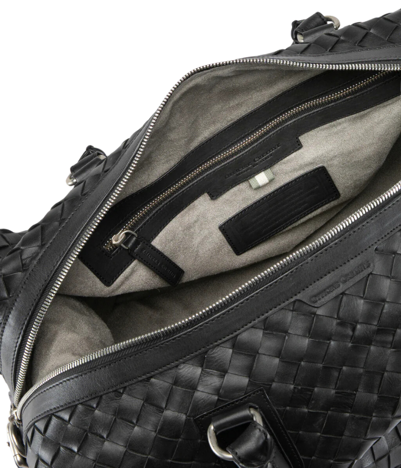 Handwoven black leather Armor 019 Travel Bag with durable handles, an adjustable shoulder strap, side buckles, and a spacious interior with a side zipper pocket. Made in Italy.