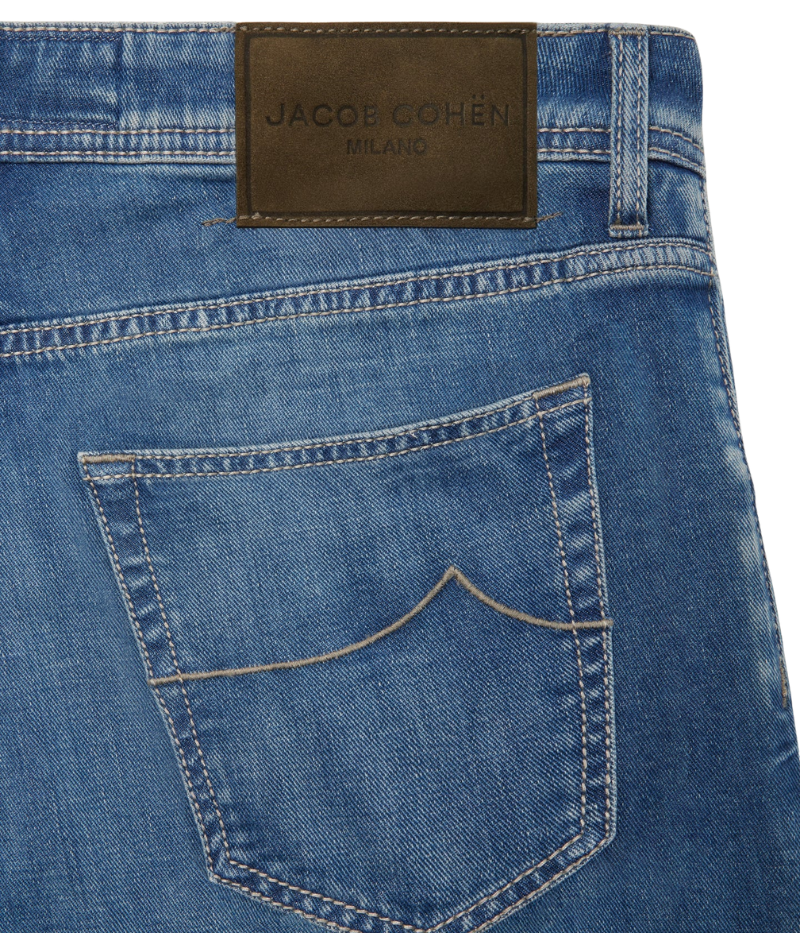 Nick 5-Pocket Jeans in medium blue stonewashed denim, showcasing their slim fit and low waist. The jeans have a lived-in feel with subtle whiskering and abrasions. Details include an olive green nubuck back patch with an engraved logo, embroidered branding, and a signature Jacob Cohën fragrance. Made in Italy from a soft stretch cotton blend.