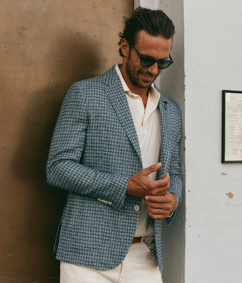 Man wearing a Lardini blue herringbone blazer with a relaxed fit, paired with a cream polo shirt and off-white trousers. The lightweight flax-wool blend fabric drapes effortlessly, featuring a subtle texture and classic herringbone pattern. Styled with sunglasses and an open collar, the look exudes effortless sophistication.