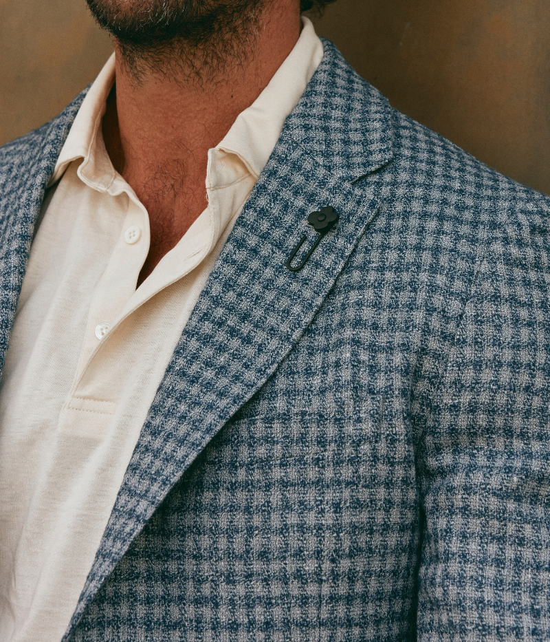 Man wearing a Lardini blue herringbone blazer with a relaxed fit, paired with a cream polo shirt and off-white trousers. The lightweight flax-wool blend fabric drapes effortlessly, featuring a subtle texture and classic herringbone pattern. Styled with sunglasses and an open collar, the look exudes effortless sophistication.