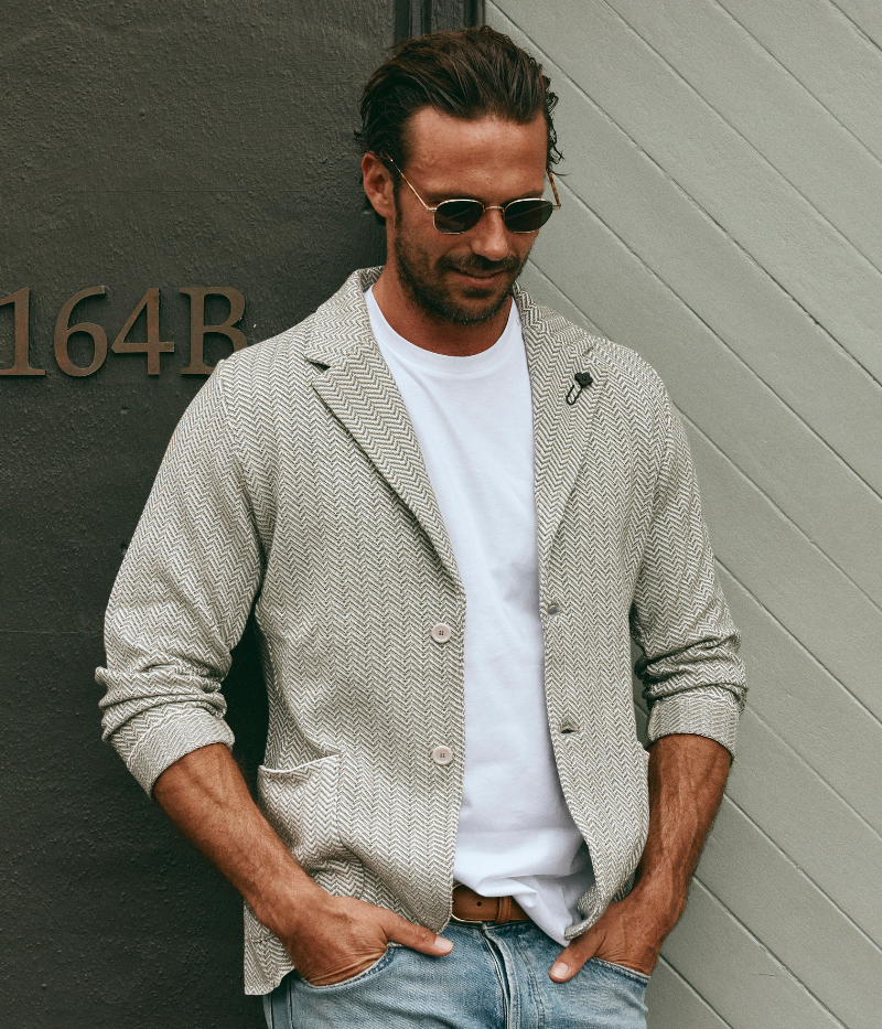 A green and white Lardini knit jacket with a herringbone pattern, soft unstructured fit, and signature floral lapel pin, made from 100% cotton, styled with a white t-shirt and jeans.