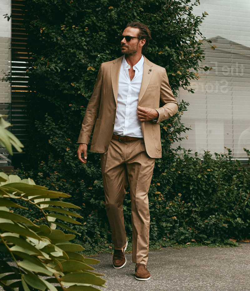 Model wearing a light brown Lardini cotton suit with a white shirt and suede loafers, featuring a single-breasted blazer with Lardini’s signature boutonnière, styled outdoors in a relaxed, modern setting.