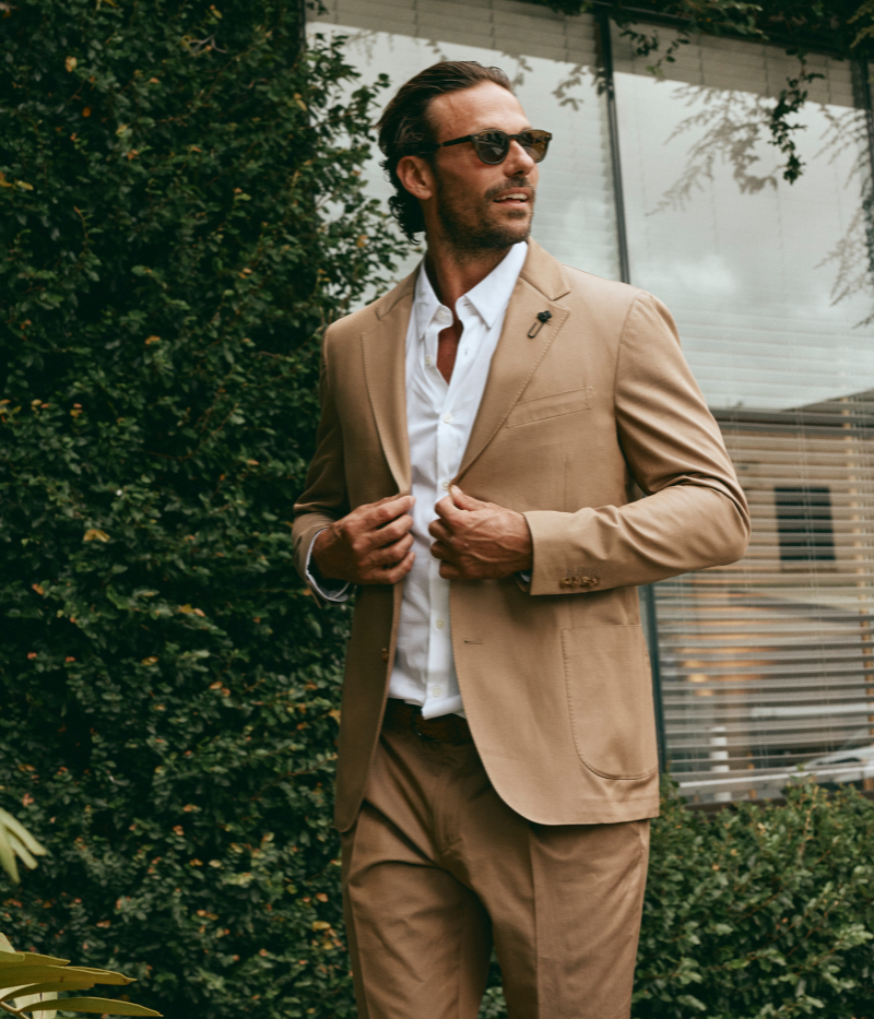 Model wearing a light brown Lardini cotton suit with a white shirt and suede loafers, featuring a single-breasted blazer with Lardini’s signature boutonnière, styled outdoors in a relaxed, modern setting.