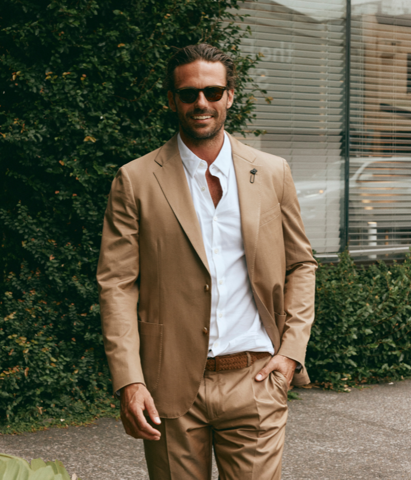 Model wearing a light brown Lardini cotton suit with a white shirt and suede loafers, featuring a single-breasted blazer with Lardini’s signature boutonnière, styled outdoors in a relaxed, modern setting.