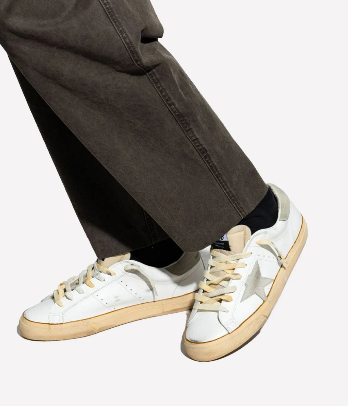 A model sitting casually, wearing Golden Goose Super Star Sneakers paired with slim-fit jeans. The sneakers’ signature distressed look and standout suede star create a relaxed, stylish feel.